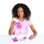 Uśmiechnięta dziewczyna z kręconymi włosami w różowej koszuli tie-dye siedzi przy białym stole, dumnie pokazując kolorową sowę Witraż na okno wykonaną z Witraż na okno, zestaw do malowania z Hazelią; inne kreacje, takie jak motyl, znajdują się przed nią.